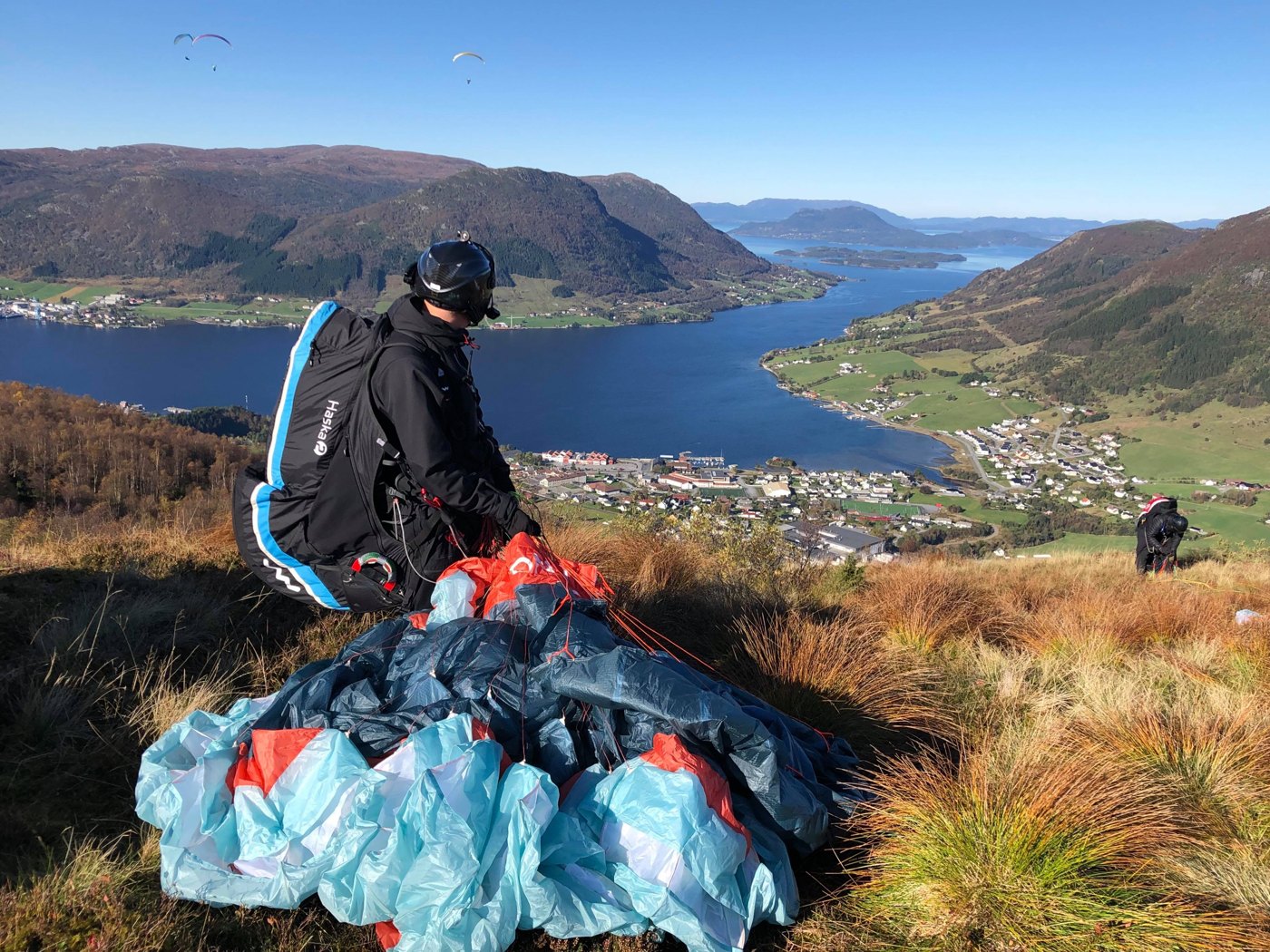 Paragliderpilot gjør klart utstyret på en fjellstart over en fjord, med andre paraglidere i luften.