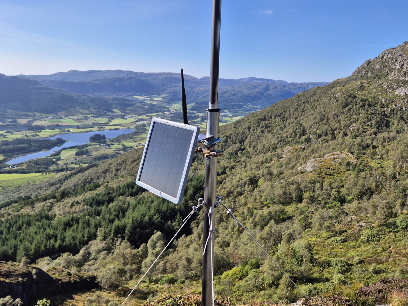 Solarnode med panel og radioantenne montert på en mast i terrenget.
